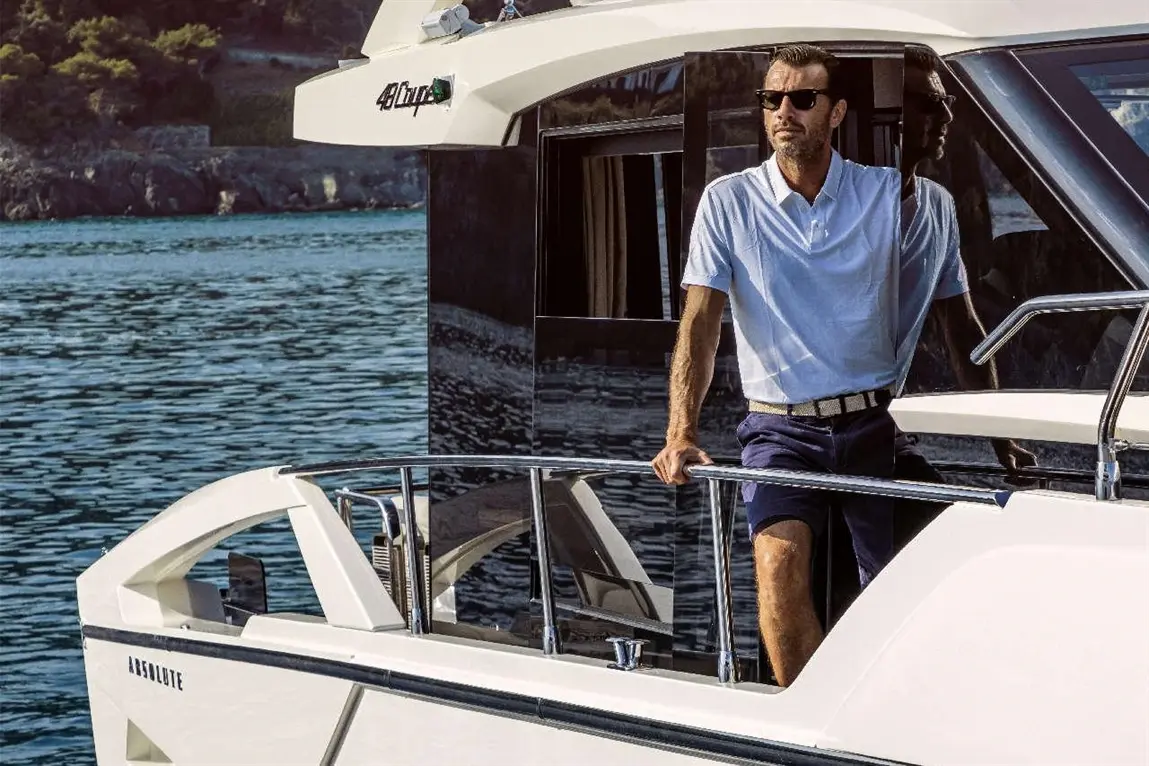 A man in sunglasses leans against the railing of a boat named 'Absolute', with calm waters in the background.